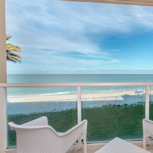 A balcony overlooking a sandy beach and calm sea, featuring two white chairs and a small triangular table, with a clear blue sky above.
