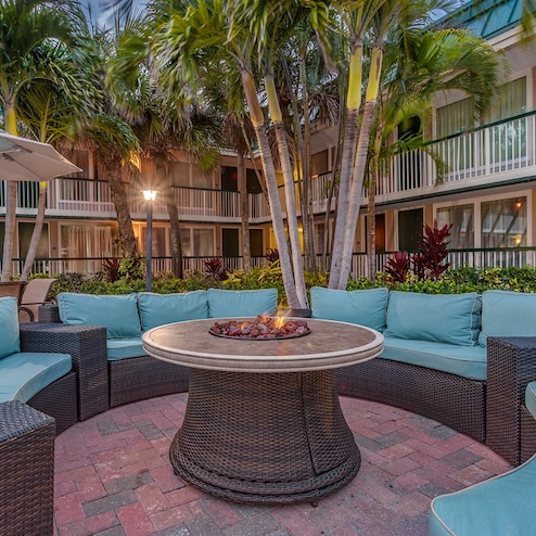 Outdoor seating area with wicker chairs and blue cushions arranged around a round fire pit, surrounded by palm trees, in front of a building with balconies at Ocean Breeze Inn Vero