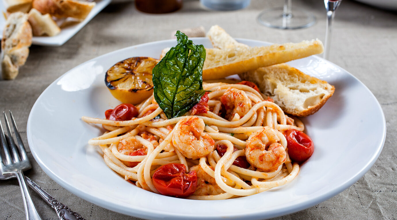 A plate of spaghetti with shrimp, cherry tomatoes, and a garnish of basil, accompanied by a breadstick and grilled lemon slice, served on a beige tablecloth with a fork and knife.
