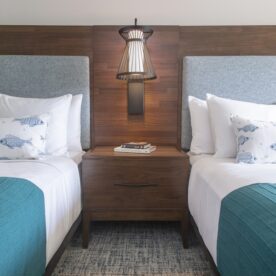 Hotel room with two queen beds featuring white linens and turquoise bed runners, centered around a wooden nightstand with a drawer and an overhead lamp.