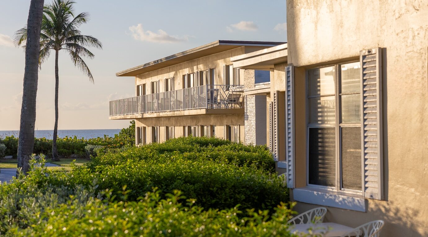 A row of buildings with balconies and shutters, set against a backdrop of palm trees and a distant ocean view during sunset.