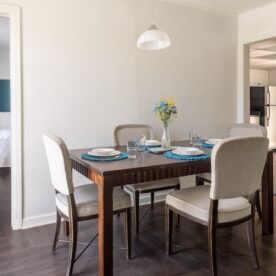 A dining room with a wooden table set for four, featuring white plates and blue placemats. There is a vase with yellow and blue flowers in the center. Four beige chairs surround the table.