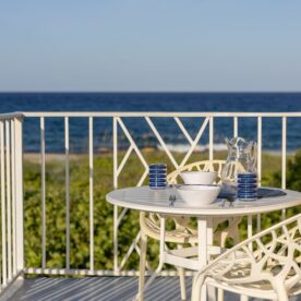 A round white patio table set with blue striped glasses, white bowls, and a glass pitcher stands on a balcony overlooking a lush landscape and the ocean under a clear blue sky.