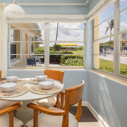 Delray Breakers on the Ocean suite. A sunlit dining area with a round table, four chairs, and four white bowls on placemats. Large windows offer a view of a building exterior, greenery, and a yellow school bus in the background.