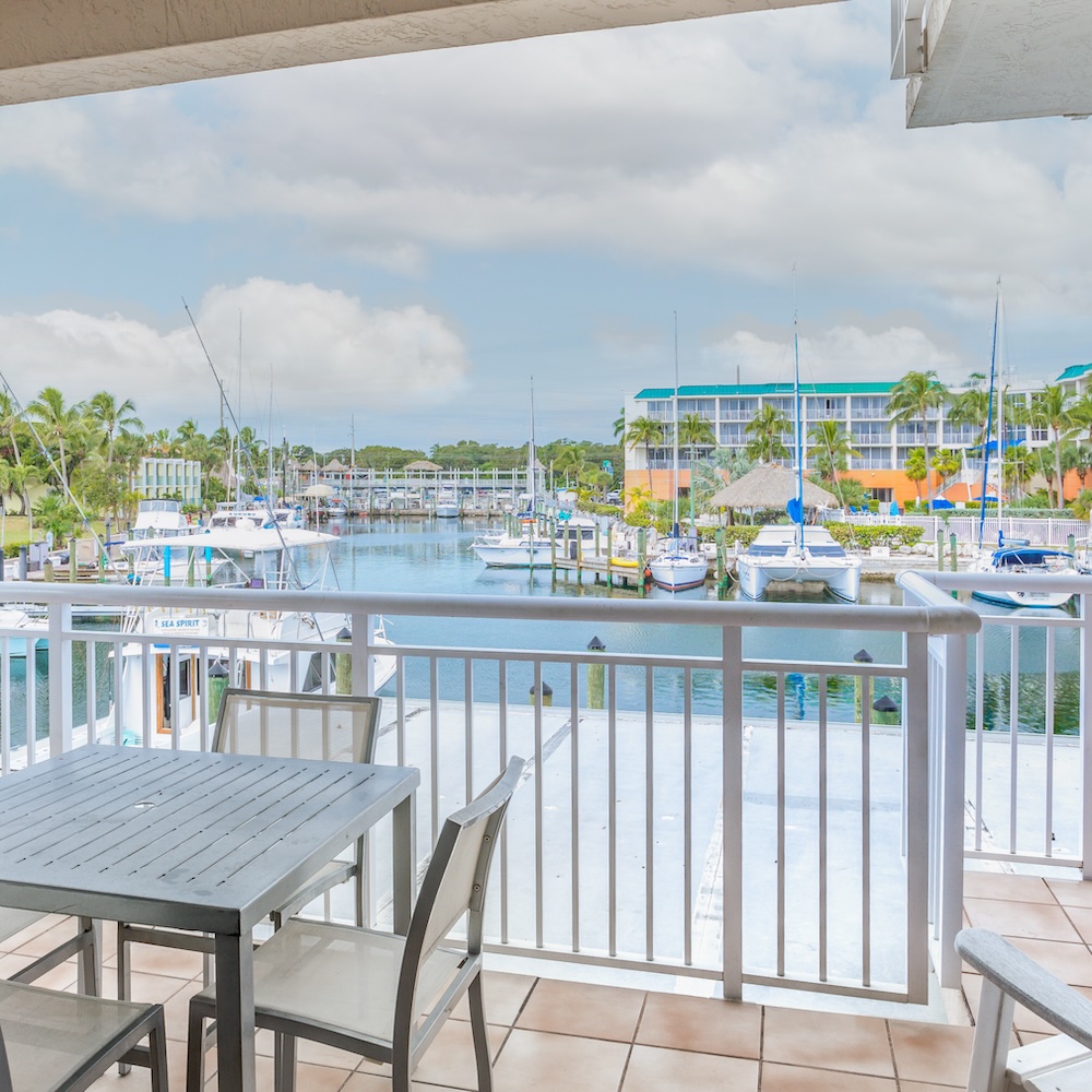 A view from Marina Del Mar's three bedroom suite balcony overlooking a marina with docked boats. The scene includes a table and chairs on the balcony, and buildings with palm trees across the water.