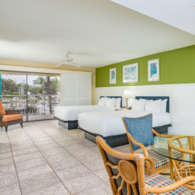 A hotel room with two double beds, a green accent wall, wicker chairs, and a table. The room has tiled flooring and a view of an outdoor area through sliding glass doors.