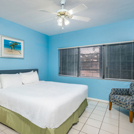 A bedroom with a light blue wall, a large bed covered with white linens, an armchair with patterned fabric, a ceiling fan, and two windows with blinds. A beach-themed picture is on the wall.