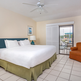 A tidy hotel room with a king-sized bed, a teal headboard, a bedside lamp, a framed wall picture, a white ceiling fan, an orange armchair, and a sliding glass door leading to a balcony with an outdoor view.