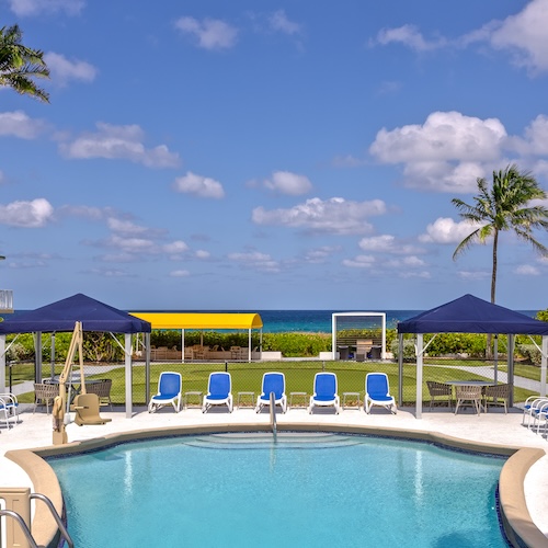 A swimming pool with blue lounge chairs in the foreground, gazebos, a lawn with seating areas, and a view of the ocean with a palm tree and blue sky with scattered clouds in the background.