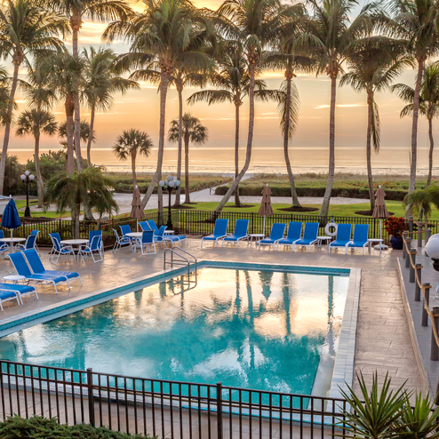 A tranquil outdoor pool area with blue lounge chairs and palm trees overlooks a sandy beach and ocean at sunset.