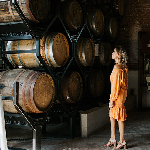 A woman in an orange dress standing in front of wine barrels.