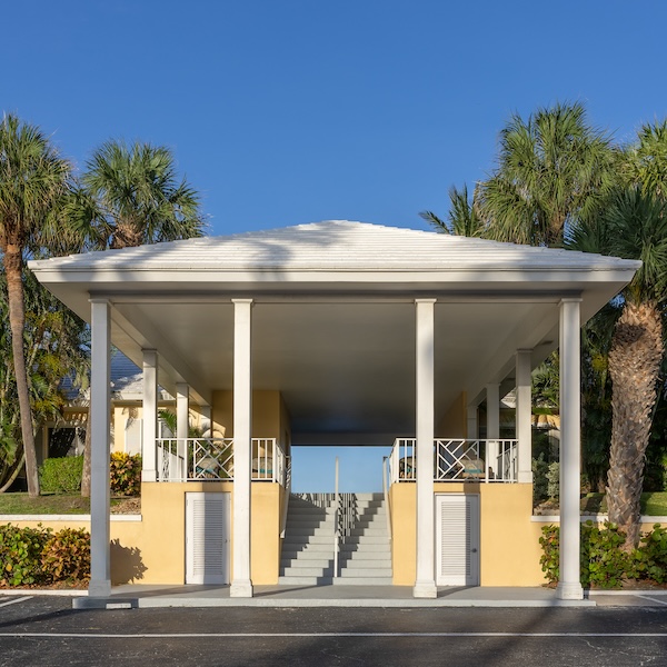 A white-pillared open structure with a staircase in the middle, yellow walls, and surrounded by palm trees under a clear blue sky.