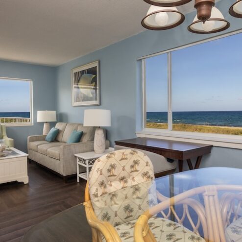 A brightly lit living and dining area with sea-themed decor, large windows overlooking the ocean, and a beige sofa beside a wooden table and chairs.