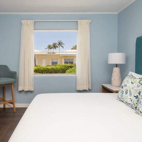 A bedroom with a double bed, blue accent chair, bedside table with a lamp, and a window with cream curtains showing an outdoor view of buildings and palm trees.