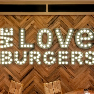 Neon sign reading "#love burgers" on a wooden slat wall in a restaurant, with tables and woven chairs in the foreground.