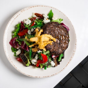 Top view of a plate with a grilled beef burger and colorful mixed salad, garnished with crispy fries, on a white surface.