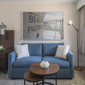 A cozy room with a blue sofa, wooden nesting coffee tables, and a "beach patrol" framed poster above the sofa. there's also a neatly made bed to the side.