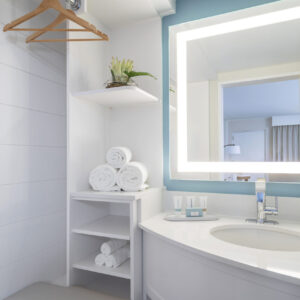 A modern bathroom featuring a backlit mirror above a sink, built-in white shelving with towels and a plant, and a reflected view of a bedroom.