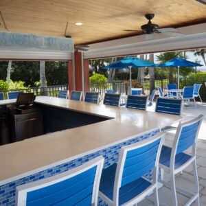 Outdoor poolside bar with blue chairs and tile detailing, named "sands 1st the bar," under a covered patio with ceiling fans and lush greenery in the background.
