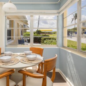 Enclosed sunroom with a round table, wicker placemats, and four bowls. Large windows offer a view of a patio, pool, and a yellow awning outside.