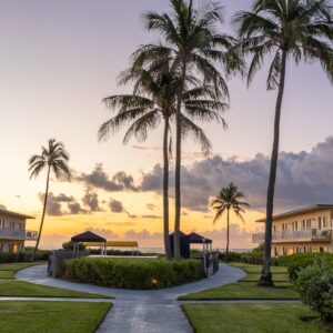 Sunset view of a resort with palm trees, manicured lawns, and pathways. Buildings flank both sides, and the ocean is visible in the background under a colorful sky.