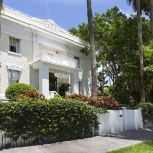 White colonial-style mansion surrounded by tropical greenery and enclosed by a white picket fence on a sunny day.