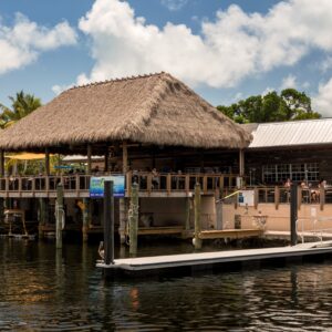 A thatched-roof tiki bar by a dock with people and boats under a partly cloudy sky.