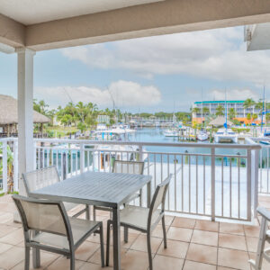View from a balcony overlooking a marina with boats and distant buildings, featuring a table and chairs in the foreground.