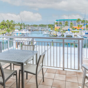 View from a balcony overlooking a marina with boats and distant buildings, featuring a table and chairs in the foreground.