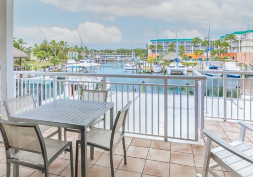 View from a balcony overlooking a marina with boats and distant buildings, featuring a table and chairs in the foreground.