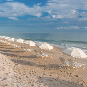 A row of white beach umbrellas and lounging chairs along an empty sandy beach with the ocean in the background on a sunny day.