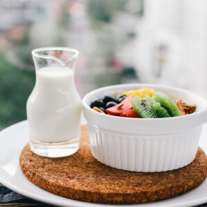 A nutritious breakfast consisting of a bowl of fruit and granola alongside a small jug of milk, set on a balcony with a blurred cityscape background.