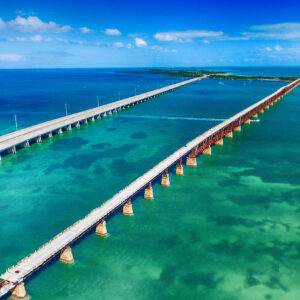Aerial view of a long bridge over clear turquoise waters, connecting two landmasses under a bright blue sky.