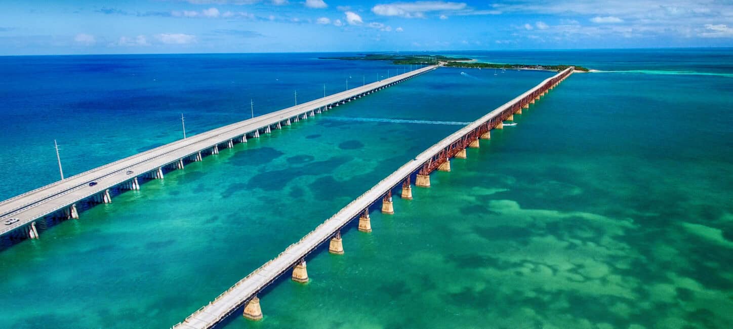Aerial view of a long bridge over clear turquoise waters, connecting two landmasses under a bright blue sky.