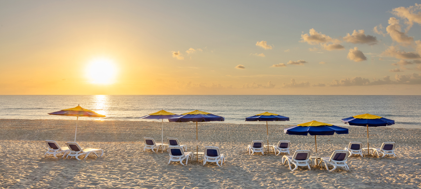 A serene beach scene at sunrise with several empty lounge chairs and yellow-and-blue umbrellas arranged in rows on the sand, facing the calm ocean.