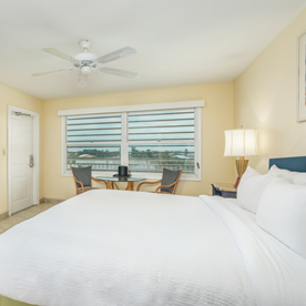 A hotel room with a large bed, nightstand, two chairs and a table by the window, a ceiling fan, and a door leading outside. The window view shows a waterfront scene.