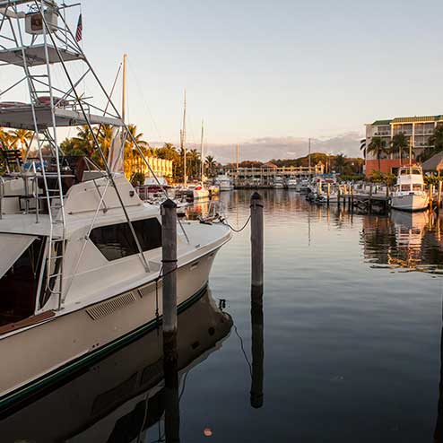 A marina at sunset with various boats docked, calm water reflecting the boats and surrounding buildings, and distant trees under a partly cloudy sky.