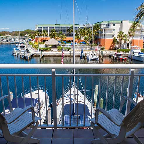 View of a marina from a balcony, featuring two white chairs in the foreground and boats docked along the water next to colorful buildings with palm trees in the background.