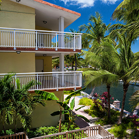 A multi-story yellow building with white balcony railings, surrounded by palm trees and tropical landscaping, overlooks a calm body of water on a sunny day.