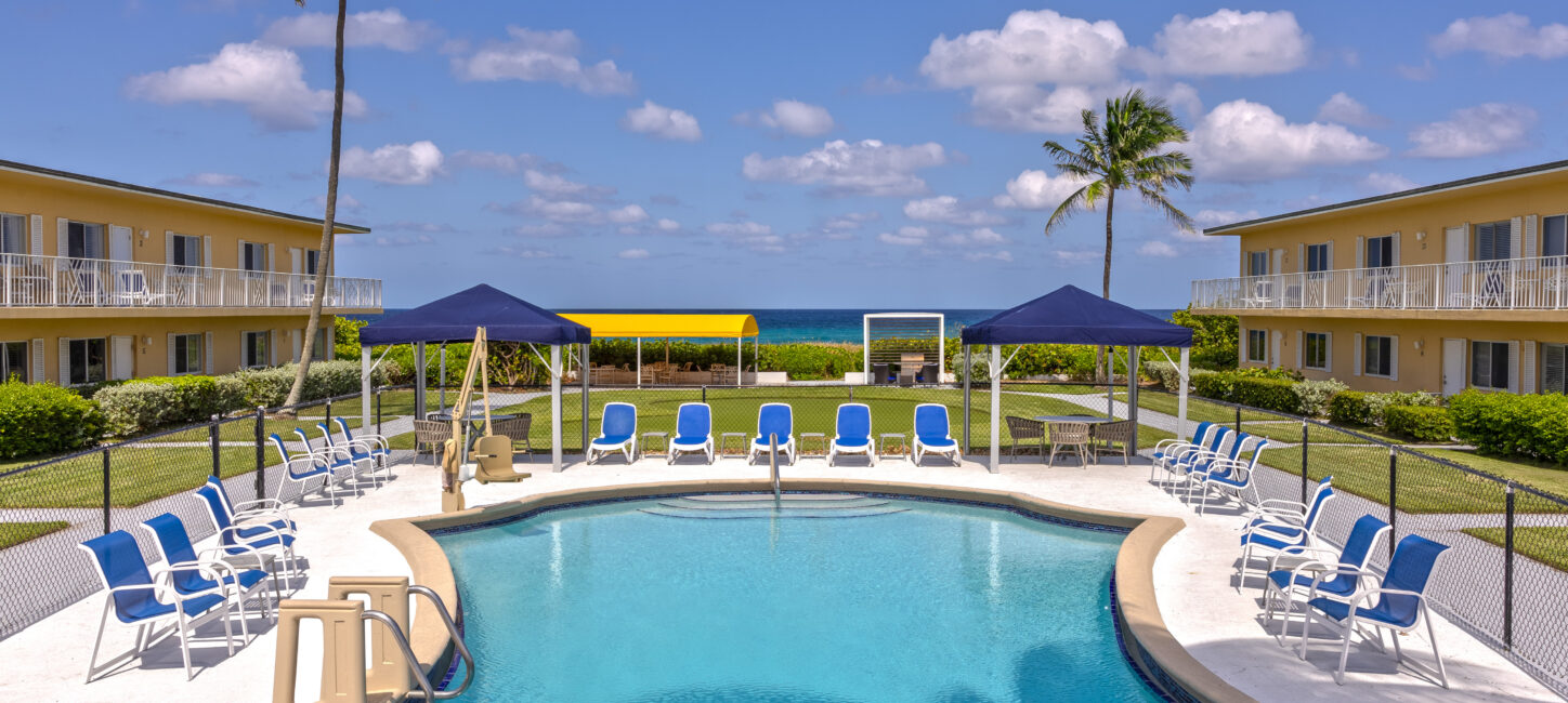 A swimming pool surrounded by lounge chairs and blue canopies, flanked by two beige buildings, with a view of the ocean in the background under a clear sky with scattered clouds.