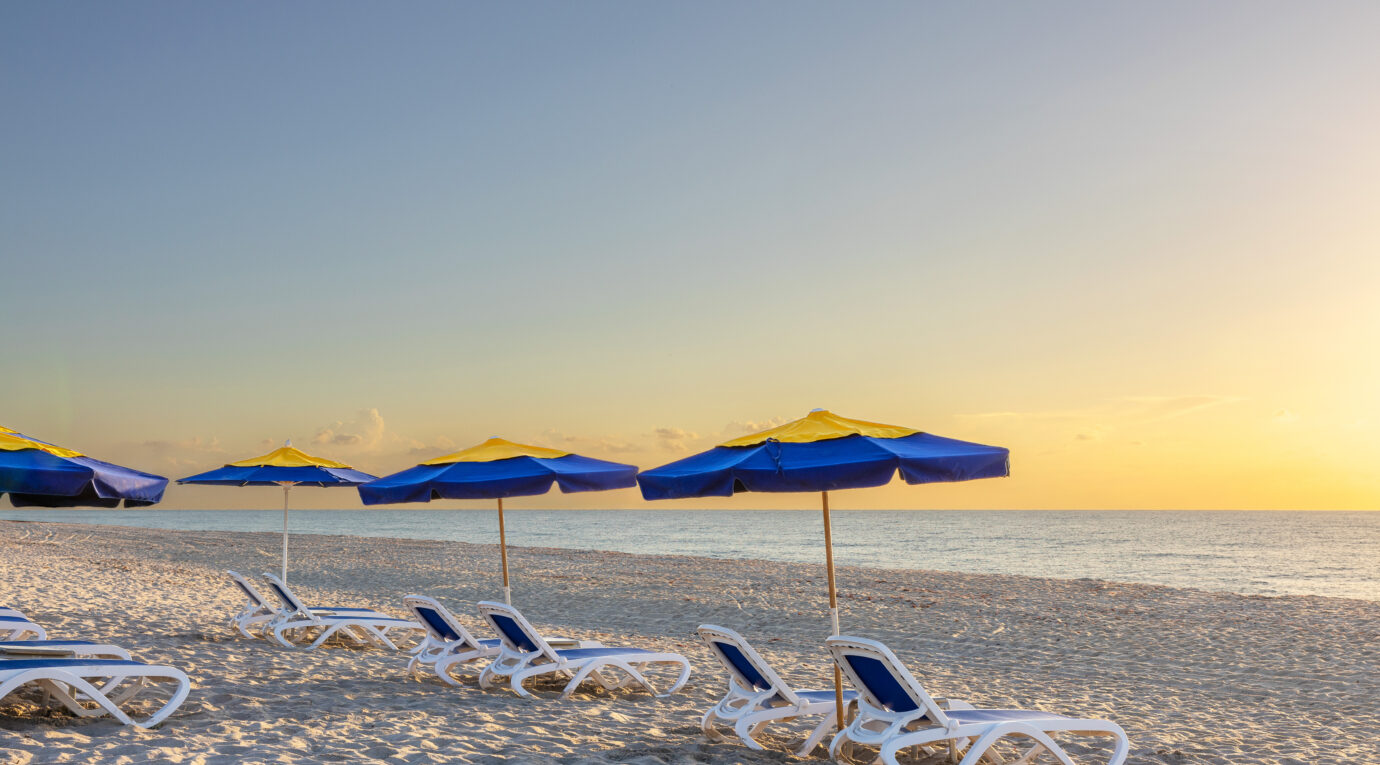 Beach scene at sunrise with empty lounge chairs and blue-and-yellow umbrellas lined up on the sandy shore facing the calm sea.