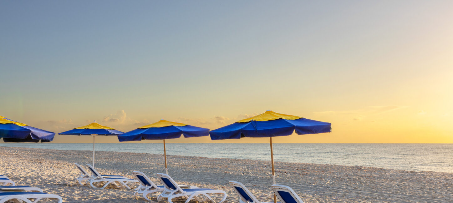 Beach scene at sunrise with empty lounge chairs and blue-and-yellow umbrellas lined up on the sandy shore facing the calm sea.