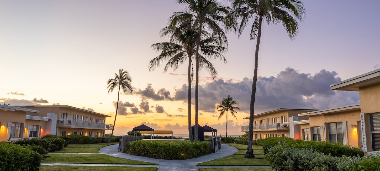 A serene resort at sunset features several two-story buildings, palm trees, and a central walkway leading to the ocean.