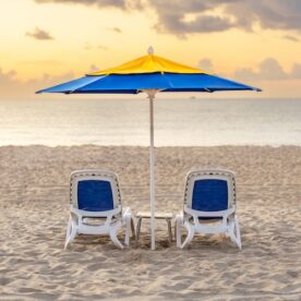 Two empty lounge chairs sit under a yellow and blue beach umbrella on a sandy beach at sunrise, facing the calm sea with a cloudy sky.