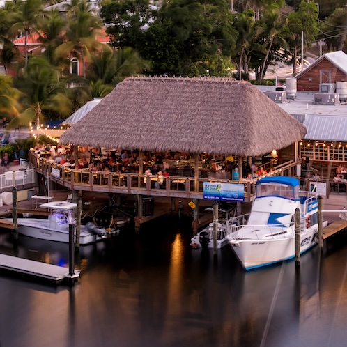 A tiki hut-style restaurant on the water, bustling with people. Boats are moored nearby. Surrounded by lush greenery and softly lit by string lights in the evening.